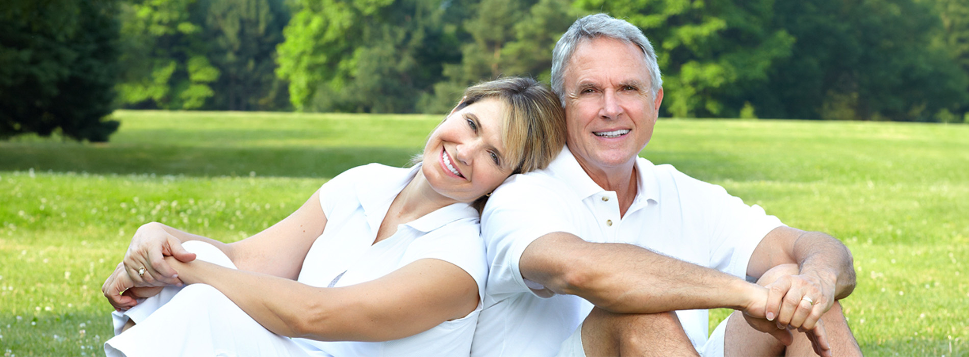 The image shows an older man and woman sitting closely together on grass, smiling at the camera, with a field behind them.